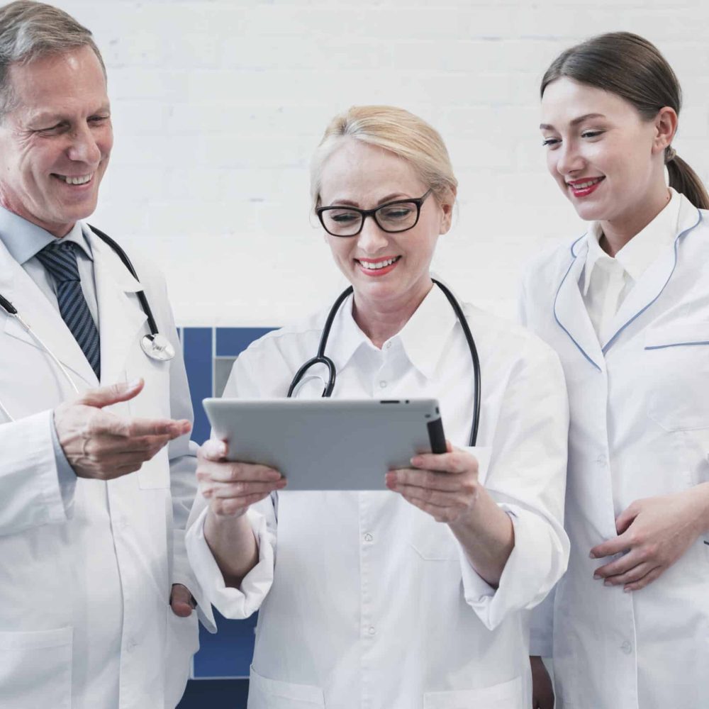 Three doctors, two women and one man, wearing white coats and stethoscopes, looking at a digital tablet and discussing in a hospital setting.